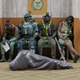 Four soldiers sit on a crowded bench in a waiting room with a bodybag on the floor in the foreground.